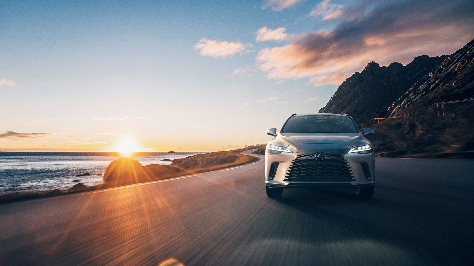 Lexus RX drives down a coastal road as the sun sets into the ocean in the background
