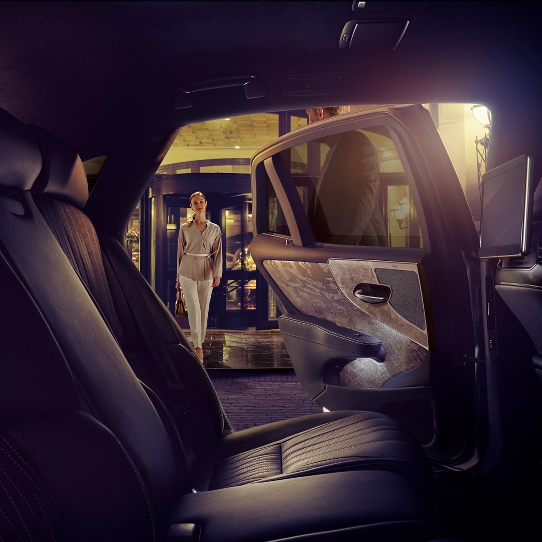 Wide shot from inside a Lexus LS, of a woman as she walks toward the LS. The back door is being held open by a man.