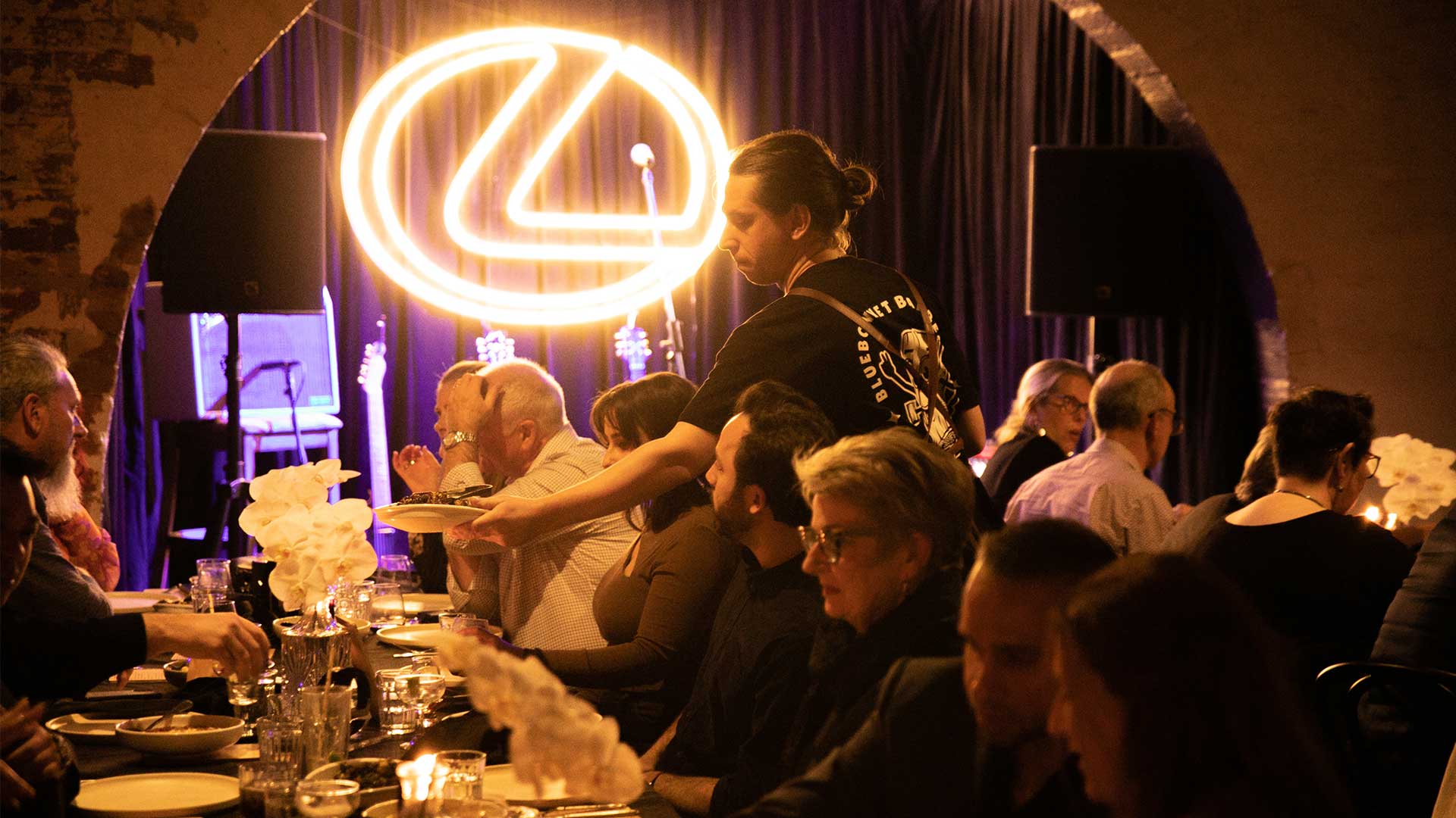 People eating dinner at a long table with a Lexus logo light in the background