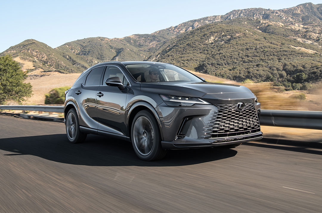 A black Lexus RX 450h+ plug-in hybrid vehicle driving along a road with hills in the background