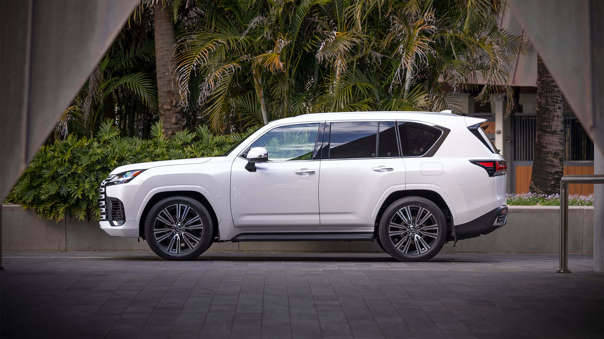 A white 2025 Lexus LX 600 Sports Luxury parked outside a modern building surrounded by trees